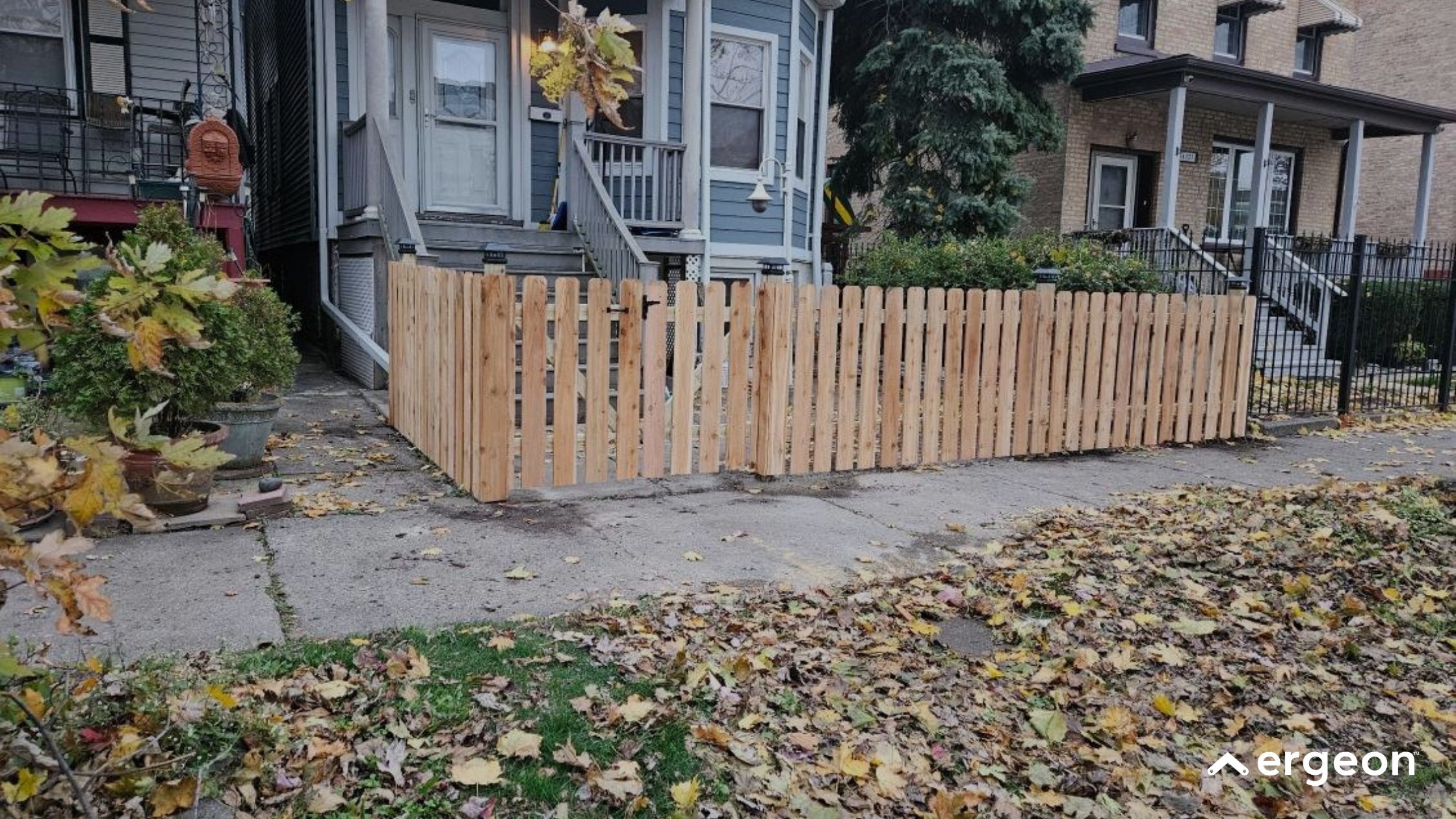 New Wooden Picket Fence Installed in Front Yard This image shows a newly installed wooden picket fence enclosing the front yard of a residential property. The fence is constructed from vertical wooden slats with evenly spaced gaps, and includes a gate with a latch at the front. The yard is situated between two neighboring houses, one painted blue and the other in a brick facade. The ground outside the fence is covered with fallen leaves, indicating an autumn setting. The fence serves both as a boundary marker and enhances the aesthetic appeal of the home's entrance.