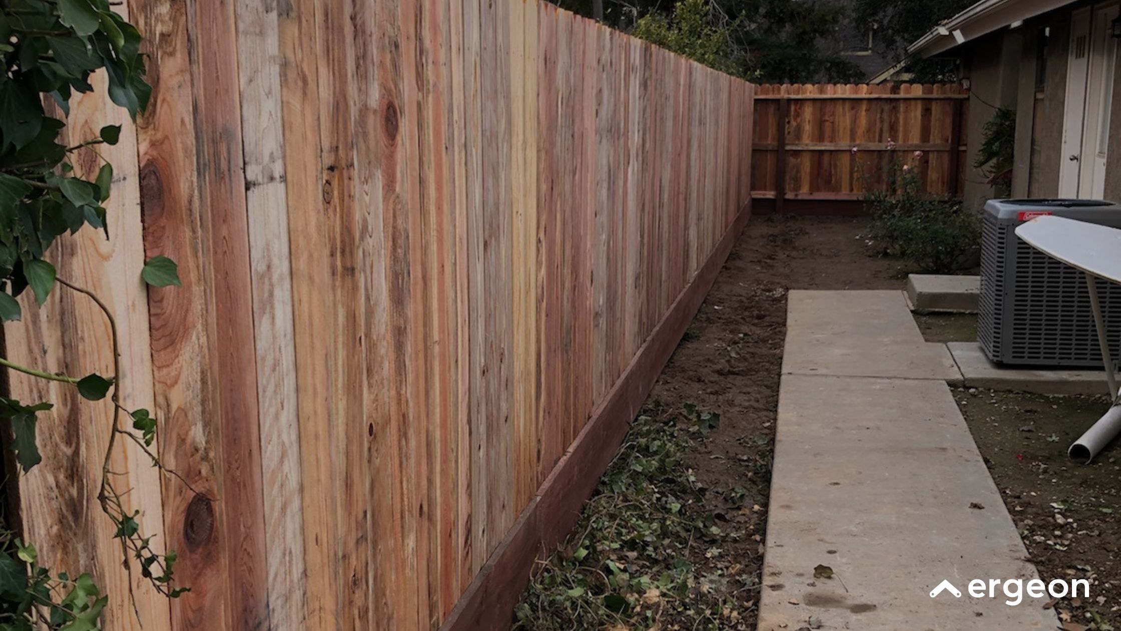 Newly Installed Wooden Privacy Fence This image showcases a freshly constructed wooden privacy fence made from vertical wooden planks with natural wood tones. The fence is installed along a narrow side yard or pathway with concrete walkways on the right side and some vegetation on the left. The scene includes elements of a residential setting, such as an HVAC unit and part of a house structure. This type of fence provides privacy and boundary definition for residential properties.