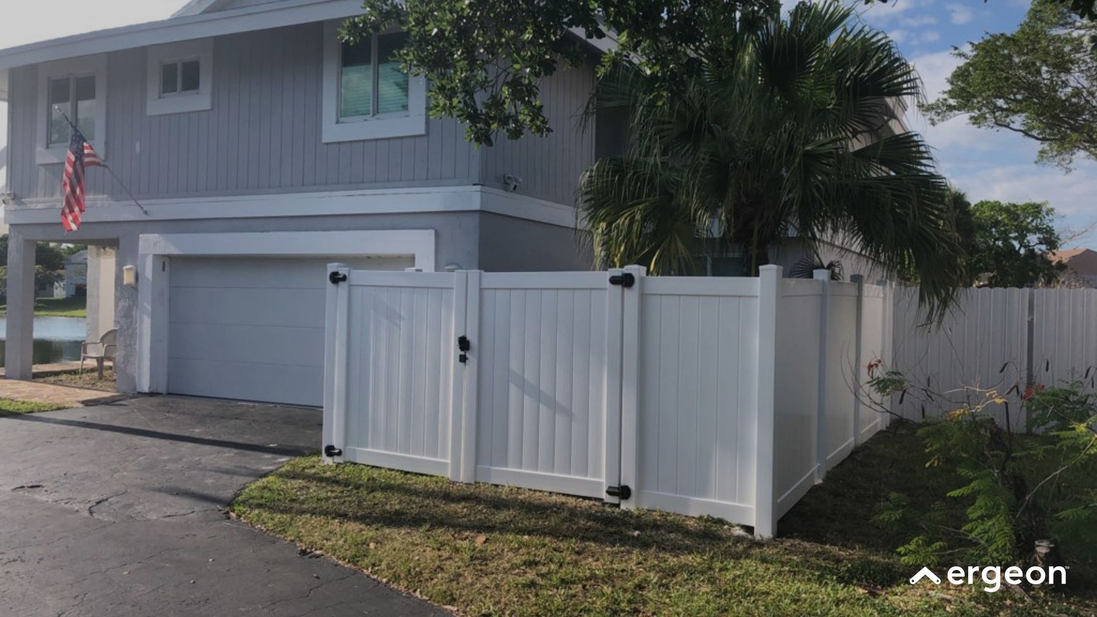 White Vinyl Fence with Double Gate Enclosing Corner of Residential Property This image depicts a corner section of a residential property enclosed by a white vinyl privacy fence featuring a double gate with black hardware. The fence surrounds a grassy area adjacent to the home's driveway and garage. The house is a two-story structure with gray siding and white trim. An American flag is hanging near the garage area. The setting appears to be a suburban neighborhood with trees and a body of water visible in the background. This type of fencing provides privacy and security while maintaining a clean, modern aesthetic suitable for residential properties.