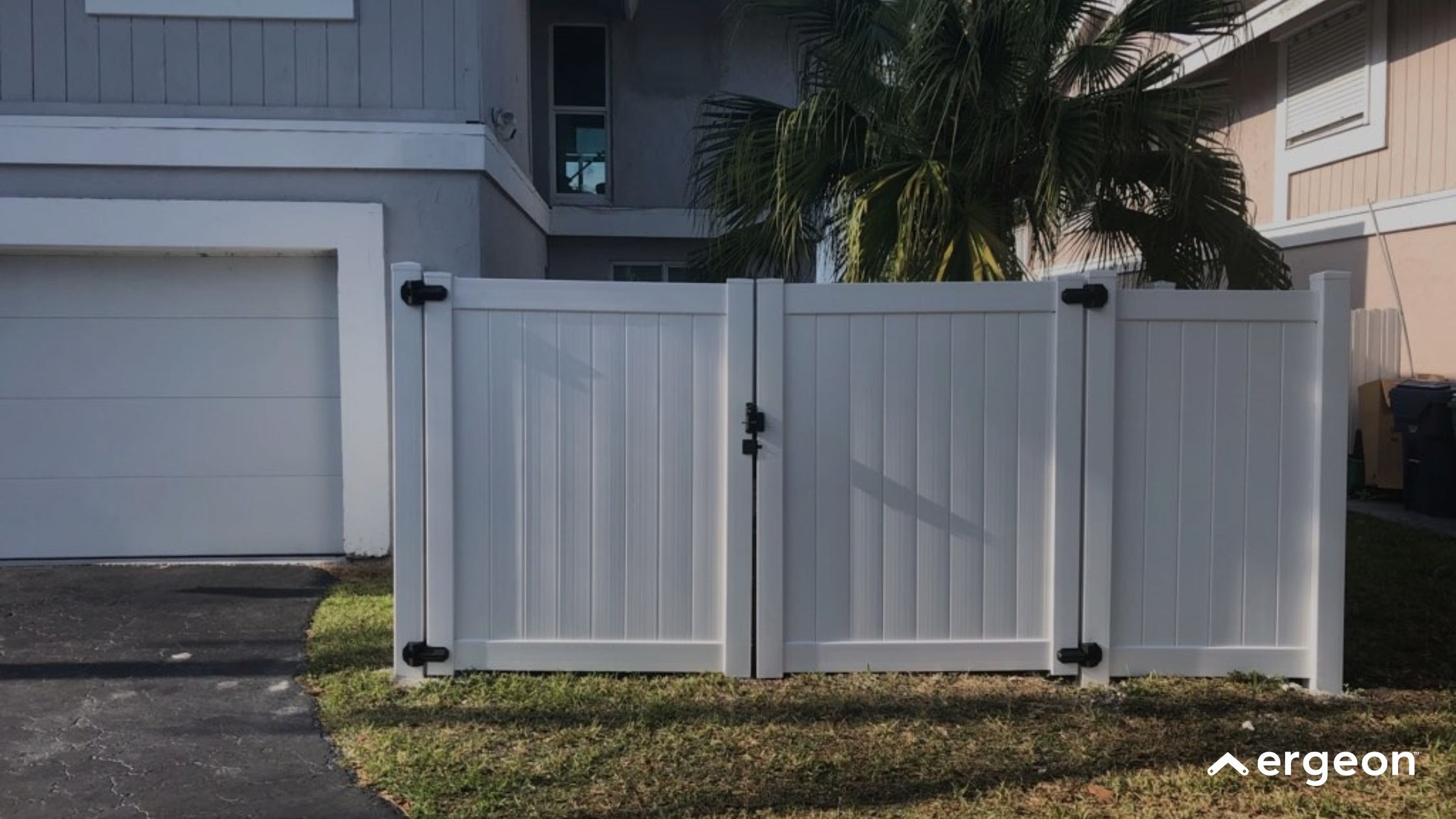 White PVC Double Gate with Black Hinges This image shows a white PVC double gate installed at the driveway entrance of a residential property. The gate panels are vertically slatted and mounted with black metal hinges and a latch mechanism for secure closing. The gate is positioned on a grassy area adjacent to a paved driveway leading to a garage door. Residential buildings and palm trees are visible in the background.