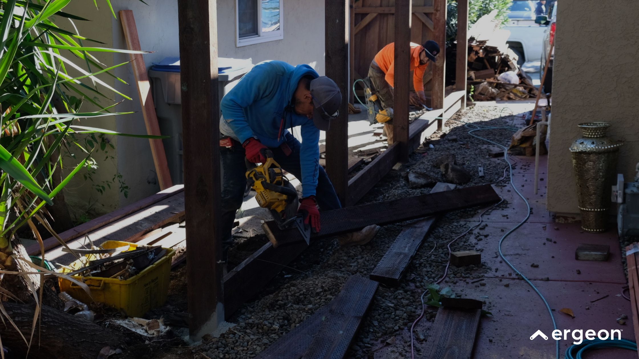Construction Workers Installing Wooden Decking Alongside Residential Building The image shows two construction workers engaged in the installation of wooden decking alongside a residential building. One worker in a blue hoodie is using a yellow power saw to cut a wooden plank, while the other worker in an orange shirt is preparing the ground or setting up the framework further along the path. The area is filled with gravel and construction materials such as wooden planks and tools. Electrical cords are visible on the ground, indicating active equipment use. This scene highlights practical outdoor residential construction work focused on decking or patio installation.
