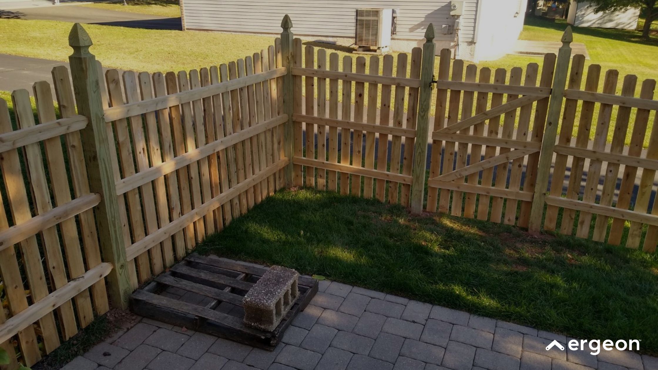 Wooden Picket Fence Corner with Gate on Patio and Lawn This image shows a newly installed wooden picket fence corner enclosing a small yard area. The fence includes vertical pickets with pointed tops, supported by horizontal rails and corner posts with decorative finials. A wooden gate with diagonal bracing is integrated into one side of the fence. The enclosed area features a tiled patio with a wooden pallet and concrete block on the ground, adjacent to a patch of green grass. The fence surrounds a residential backyard space, providing security and aesthetic boundary definition.