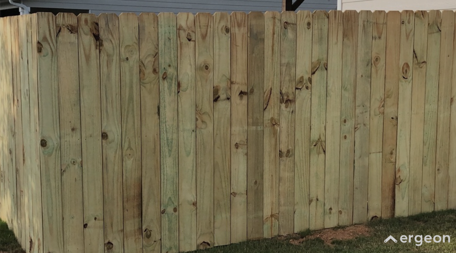 Wooden Privacy Fence Corner Installation This image shows a newly installed corner section of a wooden privacy fence made from vertical wooden planks. The fence panels are tightly aligned with visible natural wood knots and grain, providing both security and privacy for the enclosed area. The fence is situated on a grassy lawn with a house siding partially visible in the background. This type of fencing is commonly used in residential properties for boundary demarcation and privacy enhancement.