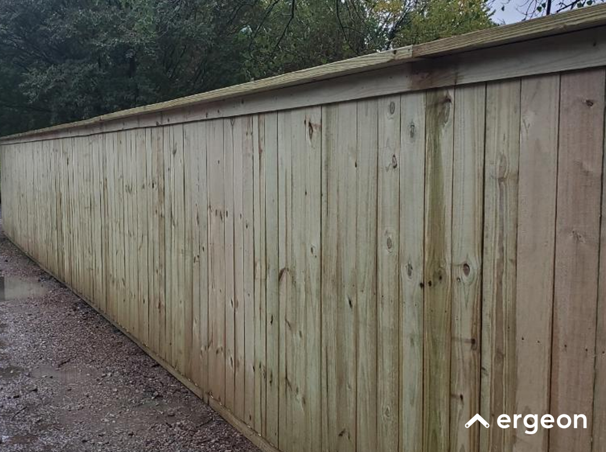 Wooden Panel Fence Exterior View This image shows a long wooden panel fence made of vertical wooden boards with a horizontal top rail. The fence is installed outdoors, adjacent to a gravel pathway, and is set against a backdrop of green trees and overcast sky. The wood appears untreated or lightly treated, with visible knots and natural grain patterns. The fence is likely used for privacy and boundary demarcation in an outdoor setting.