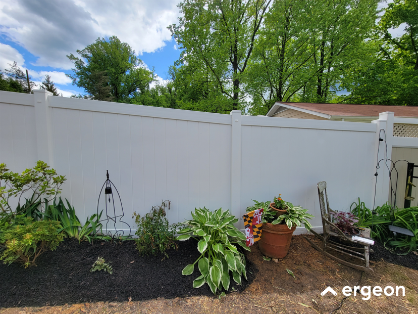 Residential Garden Area with White Privacy Fence and Decorative Plants The image shows a neat residential garden area featuring a tall white vinyl privacy fence that spans the background. Along the base of the fence is a landscaped garden bed with dark mulch and several green plants, including hostas and other foliage. There is a large terracotta pot containing a variety of plants, with a small decorative flag attached to it. To the right of the pot is a rustic, weathered wooden chair, giving a relaxed aesthetic to the garden. The scene is set in daytime under a partly cloudy sky, with mature trees visible beyond the fence. This outdoor space is suitable for residential landscaping inspiration, illustrating the use of privacy fencing, container gardening, and garden seating.