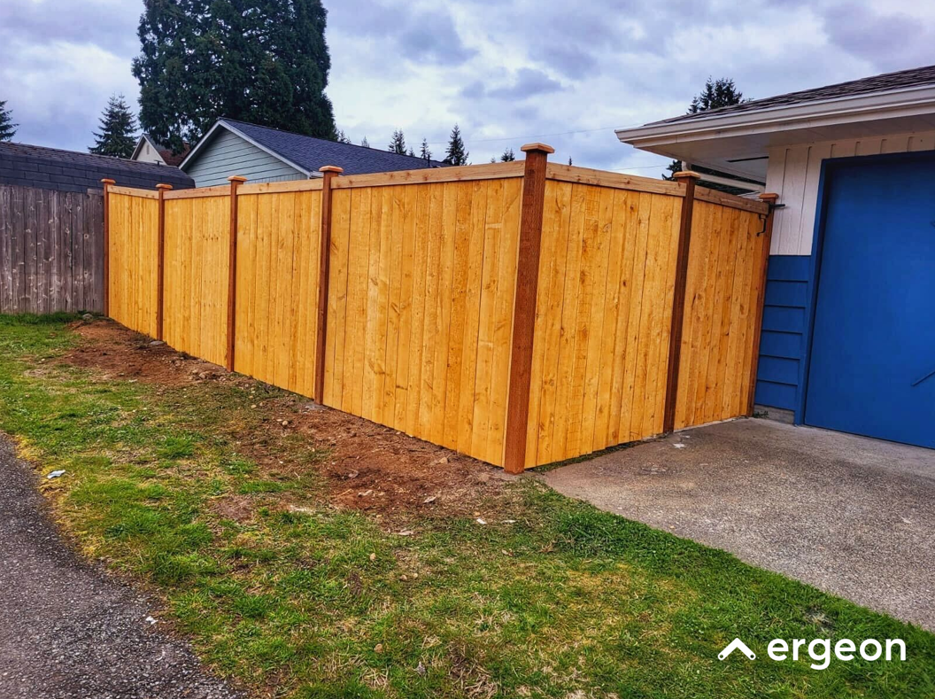 New Wooden Privacy Fence Corner Installation This image shows a recently installed wooden privacy fence forming a corner around the driveway of a residential property. The fence is constructed with vertical wooden panels supported by evenly spaced vertical posts stained a darker color. The fence borders a driveway with a concrete surface and partially encloses a grassy yard area. The background includes adjacent houses and mature trees. The photo appears to be taken on a cloudy day and includes minimal landscaping along the fence line. This type of fencing installation is suitable for enhancing privacy and security around residential properties.