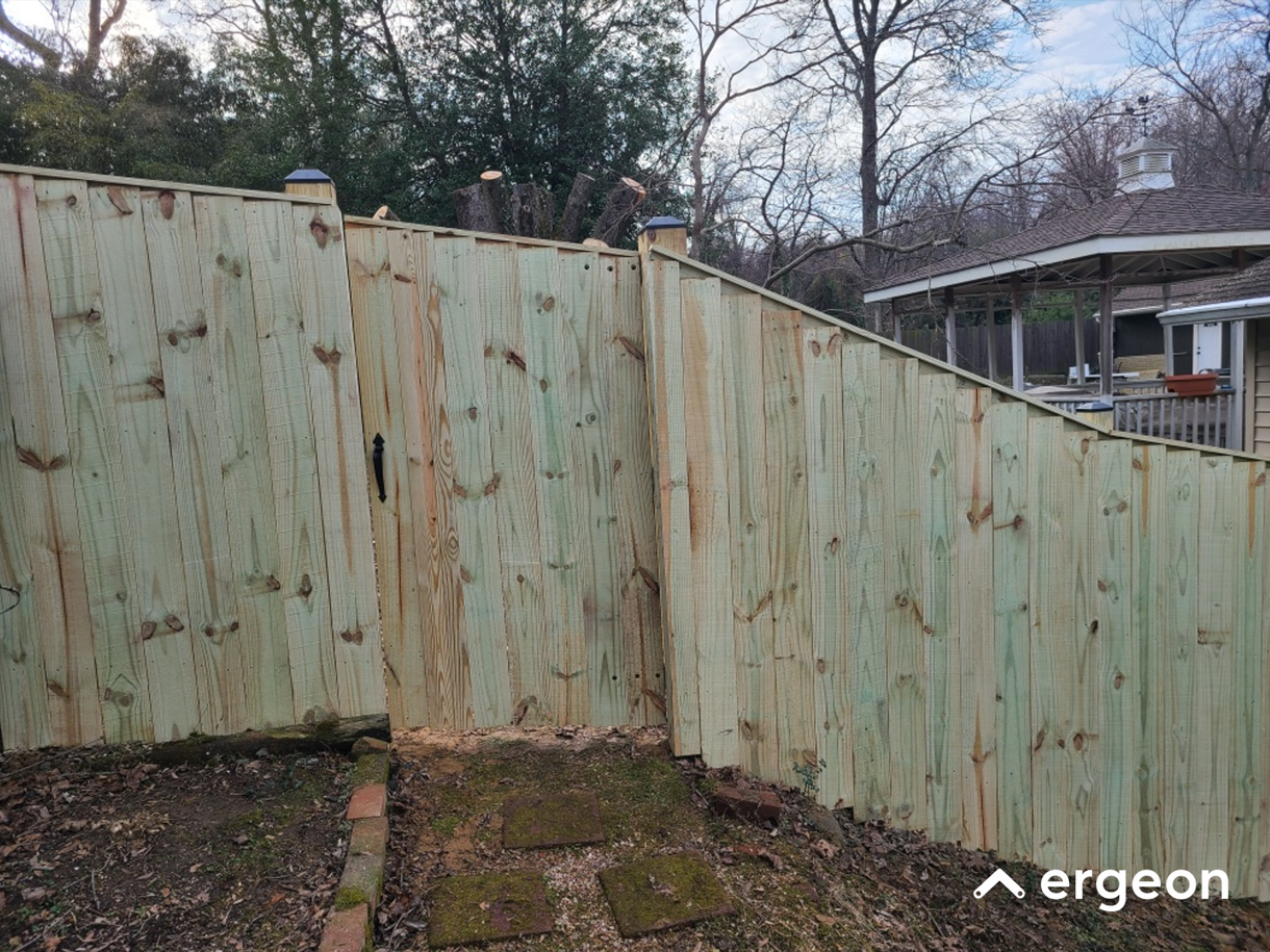 Wooden Fence Gate Installed on Sloped Yard The image depicts a newly installed wooden fence with a gate in a backyard area with a noticeable slope. The fence panels are made of untreated wood planks aligned vertically. The gate is designed with a handle and matches the style and material of the fence, blending seamlessly. Surrounding the fenced area are leafless trees, indicating a winter or early spring season. The ground in front of the gate shows a small brick border and patches of moss, with the yard slightly inclined. In the background, there is a gazebo and other residential elements visible. This image is useful for illustrating fence installation techniques on uneven terrain or slopes.