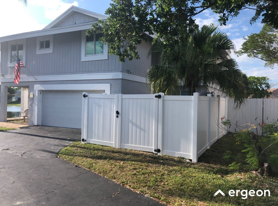 Two-Story Gray House with White Vinyl Fence and Palm Tree The image shows a modern two-story gray house with white window trims and a white two-car garage door. In front of the house, there is a white vinyl fence with black hinges and latches enclosing a small yard area. A large palm tree is situated inside the fenced yard. The house is located in a residential neighborhood with a paved driveway and a well-maintained grassy area beside the fence. The sky is clear with scattered clouds, indicating a sunny day. The image can be used to illustrate residential property exteriors featuring vinyl fencing and tropical landscape elements.