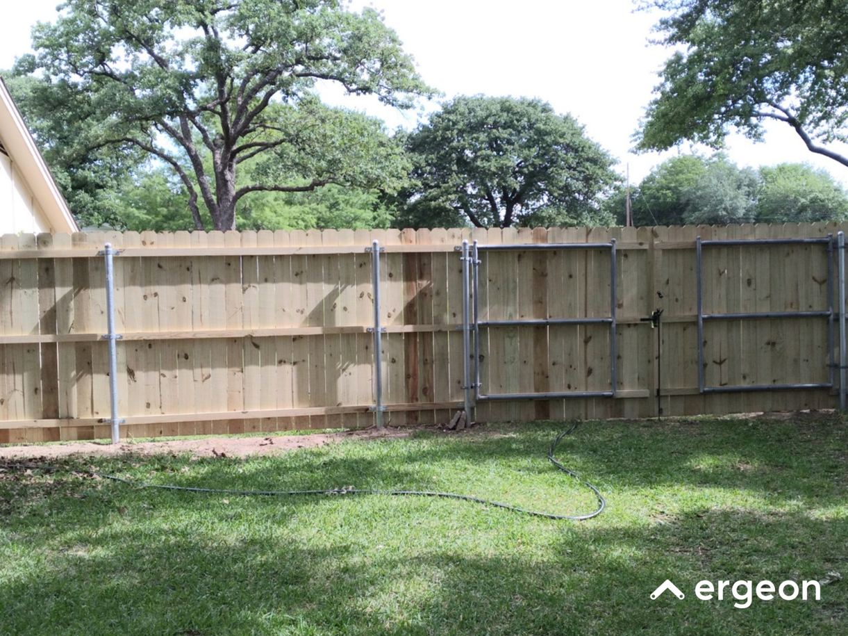Wooden Privacy Fence with Metal Gate Frame in Residential Backyard The image shows a newly constructed wooden privacy fence in a residential backyard. The fence is made of vertical wooden planks supported by horizontal braces and metal posts. Toward the right side, there is a metal gate frame attached to the wooden fence, likely for installation of a functioning gate. The yard has a well-maintained green lawn in front of the fence, and large leafy trees are visible beyond the fence line. A garden hose lies coiled on the grass. The setup is typical for providing privacy and security in residential properties.