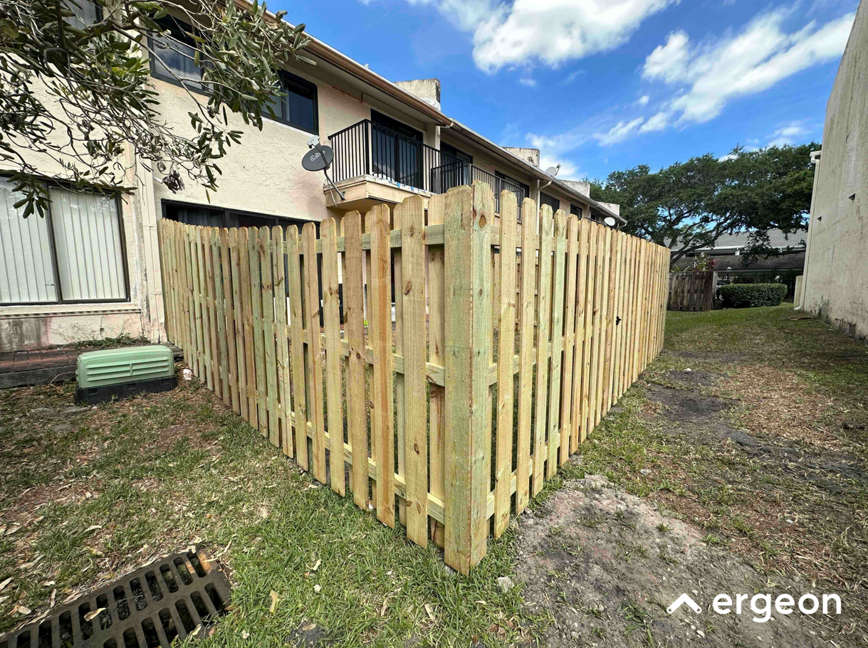 This image shows a newly installed wooden picket fence enclosing a corner of a residential backyard. The fence is constructed from light-colored wood with vertical slats and horizontal support beams, forming a secure perimeter around the backyard area adjacent to a two-story residential building. The fence includes a corner post providing structural support and extends to the inner side of the property boundary. The setting shows green grass and a drain grate near the fence, with a clear blue sky overhead. This type of fencing is typically used for privacy, security, and aesthetic improvement in residential outdoor spaces.