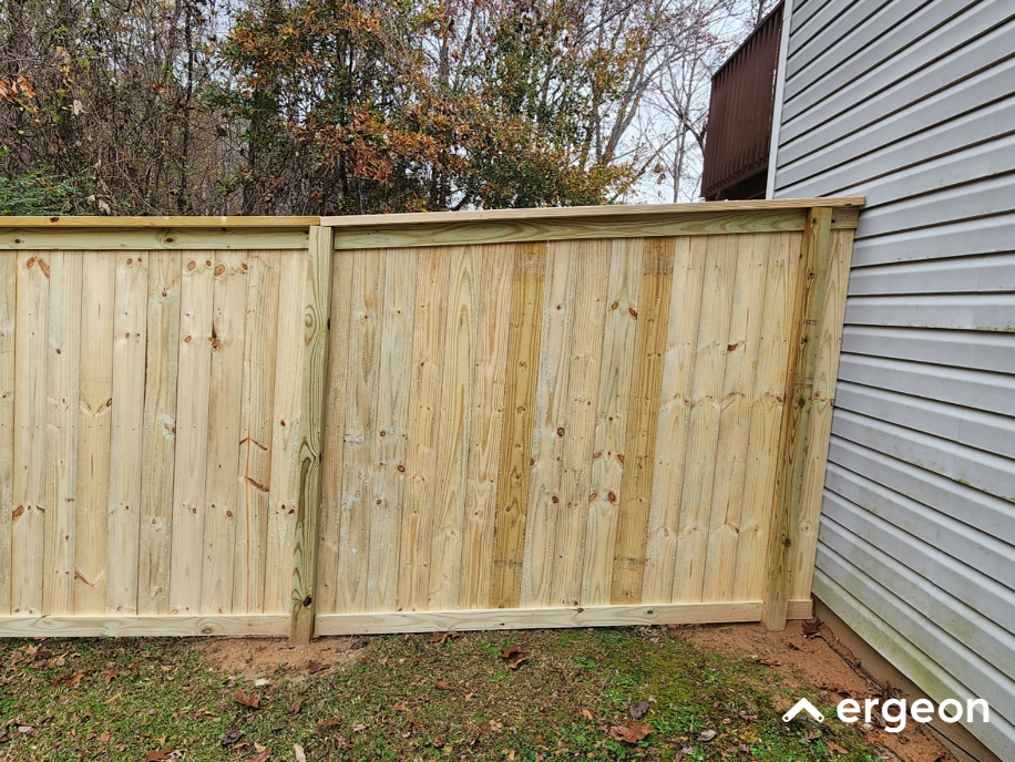 Newly Installed Wooden Fence Section Adjacent to House Exterior The image shows a newly installed section of a wooden privacy fence positioned next to the exterior wall of a house. The fence consists of vertical wooden planks secured between horizontal support beams and vertical posts. The wood appears untreated or freshly installed, with visible natural knots and grain patterns. The fence is placed on a grass and soil ground surface, providing a boundary or enclosure around the property. Trees with autumn foliage can be seen in the background, signifying an outdoor residential setting. This image can be used to illustrate fence installation techniques, types of fencing materials, or backyard boundary solutions.