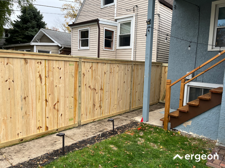 New Wooden Privacy Fence in Residential Backyard The image shows a newly constructed wooden privacy fence along the boundary of a residential backyard. The fence is made of vertical wooden planks supported by horizontal and vertical wooden beams. It borders a small concrete walkway and a grassy area adjacent to a blue house with wooden stairs leading up to a door. The backyard landscaping includes small solar pathway lights along the walkway, and the surroundings include neighboring houses in the background. This type of fence is typically used for privacy and property demarcation in residential settings.