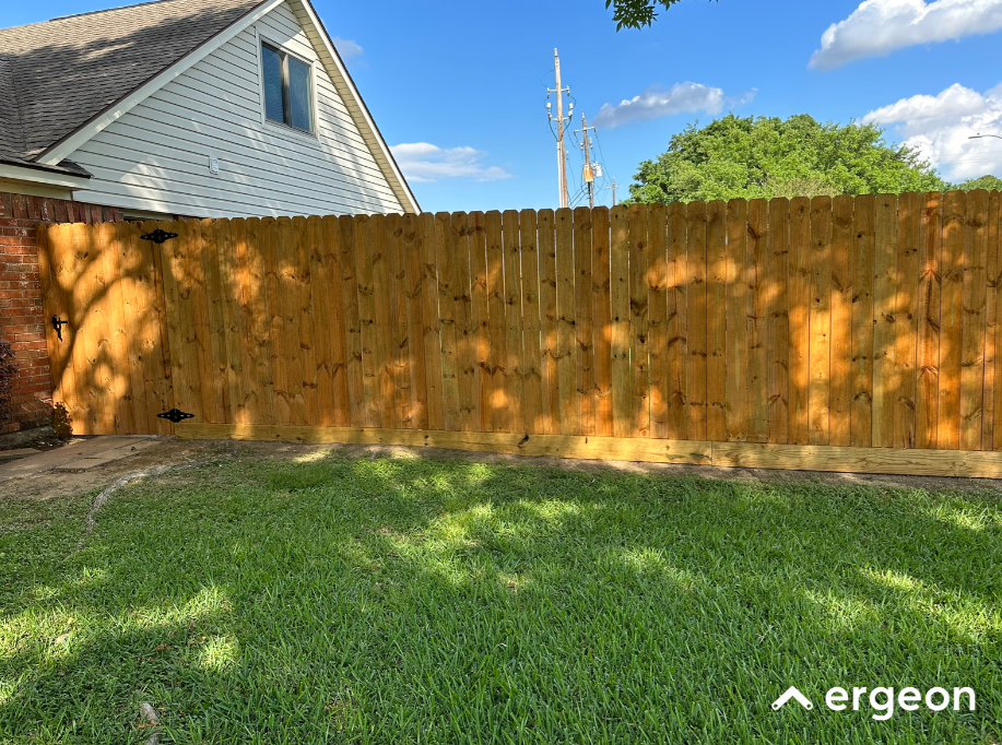 Wooden Privacy Fence in Residential Backyard This image shows a new wooden privacy fence installed in a residential backyard. The fence is made of vertical wooden planks supported by horizontal boards at the base and secured with large black metal hinges and a latch on the left side, indicating a gate. The fence runs along the property line beside a house with siding and a shingled roof visible. The yard has well-maintained green grass and some trees casting shadows on the fence. The clear sky, utility poles, and a large tree are also visible beyond the fence. This image can be used for documentation or knowledge sharing about residential fence installation, privacy fencing design, or landscaping.