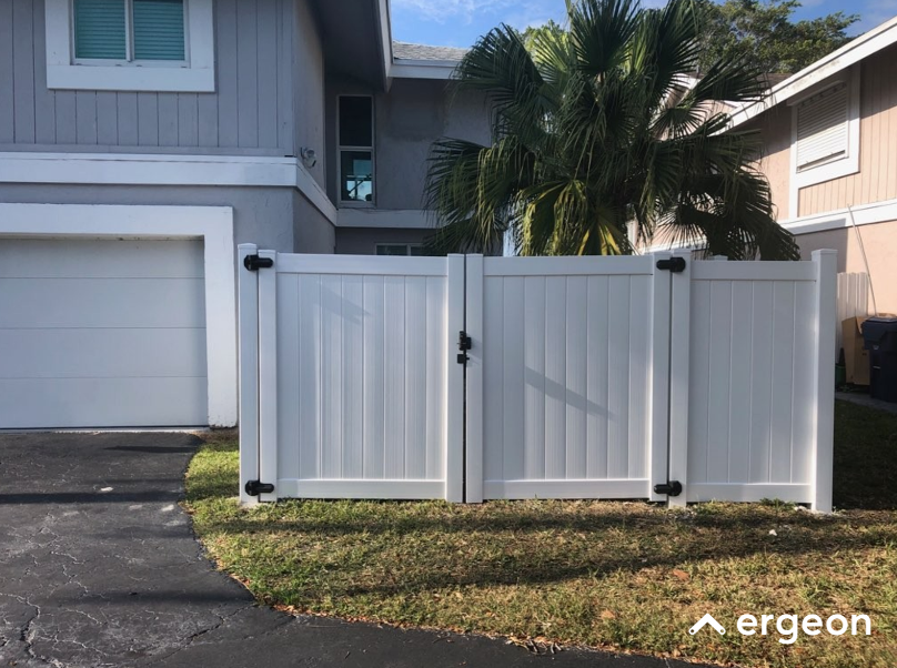White Vinyl Double Gate with Black Hinges and Lock in Residential Driveway This image shows a white vinyl double gate installed in the driveway area of a residential property. The gate features vertical paneling and is secured with black metal hinges and a central locking mechanism. It is set against a backdrop of a gray house with a garage door on the left and palm trees in the background, illustrating a typical suburban home entrance setup. The gate provides both privacy and security while complementing the exterior aesthetic of the property.