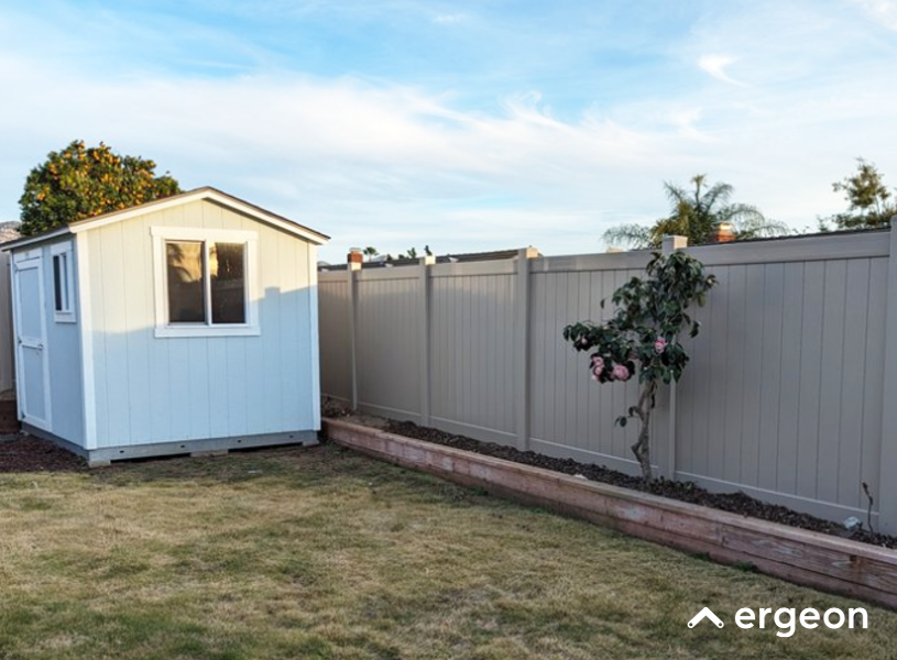 Backyard Storage Shed with Garden Fence and Raised Flower Bed This image shows a backyard scene featuring a small, light blue storage shed with a white-framed window and door located near a beige privacy fence. Adjacent to the fence is a wooden raised flower bed with soil, containing a flowering shrub with pink blossoms. The yard has a grassy area that appears partially dry or dormant, under a mostly clear sky. This setup is typical for organized residential outdoor spaces offering storage, privacy, and garden cultivation.