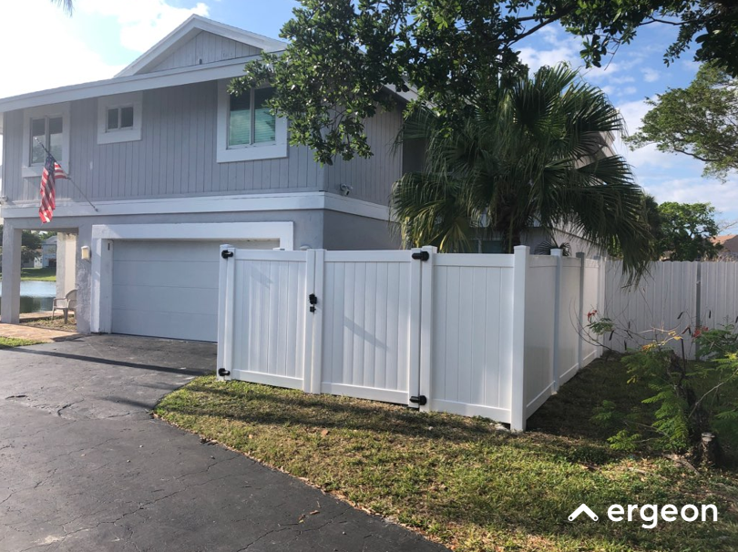 Residential Home Featuring White Vinyl Privacy Fence and Attached Garage This image shows a two-story residential house with a light gray exterior and white trim. The house features a white, double garage door and a white vinyl privacy fence enclosing the side yard. The fence includes a gate secured with black hardware. In front of the house, there is a driveway paved in asphalt and a small lawn area. The property has mature landscaping with a palm tree and other foliage visible. An American flag is mounted near the garage. The setting appears to be suburban, with clear weather and blue skies. This image can be used for documentation of residential fencing options, home exterior aesthetics, and property boundary delineation.