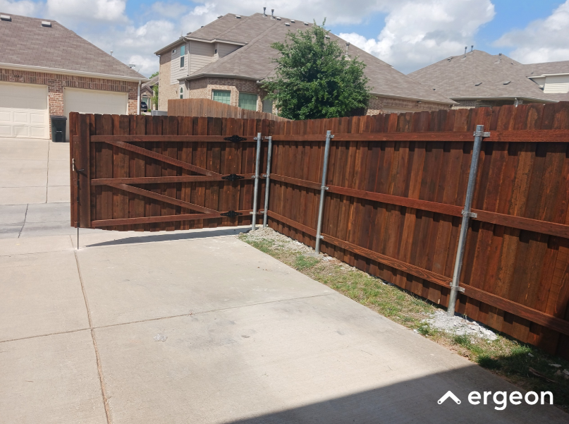 Wooden Fence with Double Swing Gate on Concrete Driveway This image shows a wooden fence with a double swing gate installed on a concrete driveway. The fence is constructed with vertical wooden planks supported by horizontal wooden beams and reinforced with vertical metal poles for added stability. The double gate features diagonal wooden braces and black metal hinges, allowing it to swing open. The setting is a residential area with several houses visible in the background, and the gate provides access between the driveway and the enclosed backyard or side yard. This type of gate and fencing is commonly used for privacy, security, and aesthetic enhancement in suburban homes.