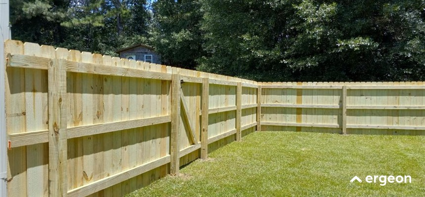 The image shows the interior side of a newly installed wooden privacy fence enclosing a backyard area. The fence is constructed from light-colored wood with vertical planks supported by horizontal rails and vertical posts for stability. The grass in the fenced area is green and well-maintained, and there is a natural wooded background outside the fence. This image can be used for documentation related to residential fencing installations, landscape design, and property boundary delineation.