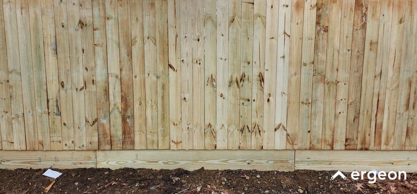 The image shows a close-up section of a vertical wooden fence installed along a garden or yard border. The fence is made of light, natural-colored wooden planks with visible knots and grain patterns. The base of the fence meets the soil ground, indicating it is outdoor fencing, likely used to provide privacy or boundary definition in residential areas.