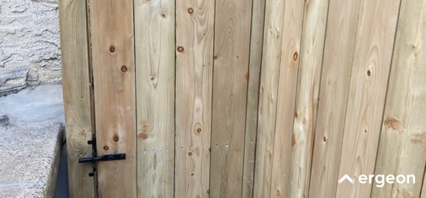 The image shows a close-up view of a wooden gate constructed from vertical wooden planks. The gate features a simple black metal latch for securing it. It is installed next to a stone wall with a concrete step visible on the left side. The wood appears to be treated or new, showcasing natural grain and knots. This gate setup is suitable for outdoor residential use, providing access control and privacy.
