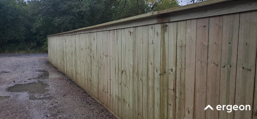 This image shows a long wooden privacy fence constructed with vertical planks, installed alongside a gravel pathway. The fence appears to be in a natural outdoor setting with trees visible in the background. The wood shows natural grain and knots, suggesting untreated or lightly finished timber. The fence provides privacy and boundary demarcation, suitable for residential or commercial use. The gravel pathway has some wet patches, indicating recent rain or water exposure. The image includes the 'ergeon' logo in the bottom right corner, indicating it could be used in the context of construction permits, fencing regulations, or property boundary guidelines.