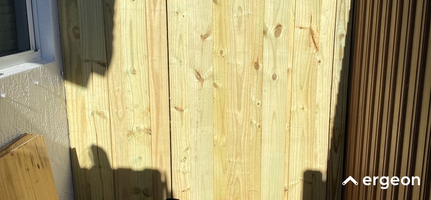 This image shows a close-up view of a wooden fence panel made of vertically aligned light-colored wooden planks. The wood appears to be newly installed or well-maintained, suggesting durability. The fence is located outdoors next to a textured wall and a window, indicating residential use. This type of wooden fencing is suitable for weather conditions such as those found in Daytona Beach, Florida, implying it may be treated for resistance against moisture, sun exposure, and wind.