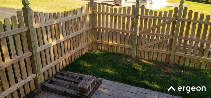 This image shows a newly installed wooden picket fence forming a corner around a grassy yard area adjacent to a paved patio. The fence is made of vertical wooden pickets supported by horizontal rails and sturdy corner posts. A wooden pallet with a concrete block is placed on the patio near the fence. The setting appears residential, demonstrating typical fence construction and installation quality suitable for outdoor boundary definition and privacy.