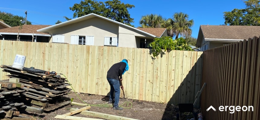 The image shows a person installing a new wooden fence in the backyard of a residential property in Lakeland. The person is working with tools on the ground near a newly constructed fence panel made of untreated wooden planks. On the left side, there is a stack of old or unused wooden materials, and on the right side, a brown fence is already in place. The background includes neighboring houses and trees, indicating a suburban setting. This image can be used to illustrate local fence installation practices and considerations for residential permit requirements in Lakeland.