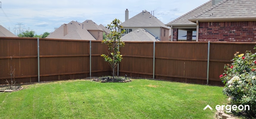 This image depicts a suburban backyard enclosed by a tall wooden privacy fence. The fence consists of vertical wooden panels supported by metal posts, providing both security and privacy. Inside the yard, there is a well-maintained grassy lawn with some planted areas, including small trees and shrubs. The background shows neighboring houses with similar roofing styles, indicating a residential neighborhood. This type of fencing is commonly used for privacy and boundary definition in residential properties.