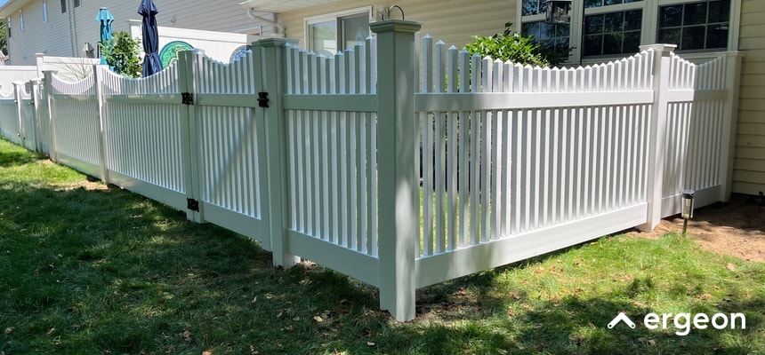 This image shows a newly installed white vinyl picket fence with vertical slats and a decorative arched design on the gate. The fence encloses a residential backyard area, including a gate with black hinges and latch hardware. The fence provides both aesthetic appeal and security for the property. It is set up on a grassy lawn adjacent to a house with windows and siding visible in the background. The fencing material appears durable and low-maintenance, suitable for creating a defined boundary in suburban home landscaping.