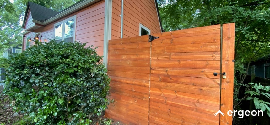 This image shows a solid wooden privacy fence attached to the side of a residential house. The fence is composed of horizontal wooden planks with a warm stained finish, secured with black metal hinges and latches. The fence provides privacy and security for the property, blending aesthetically with the home's exterior. Lush green foliage surrounds the area, indicating the fence's suitability for outdoor residential environments. The fence design suggests durability and weather resistance, likely appropriate for diverse weather conditions.