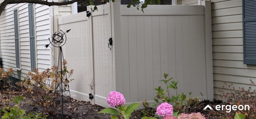This image shows a light-colored privacy fence with an integrated gate, installed adjacent to a residential house. The fence is designed with vertical panels and black metal hinges and latch for the gate. Surrounding the fence is a garden area featuring ornamental plants and blooming flowers, contributing to the curb appeal of the home. The fence provides a private enclosed space and adds to the aesthetic value of the property.