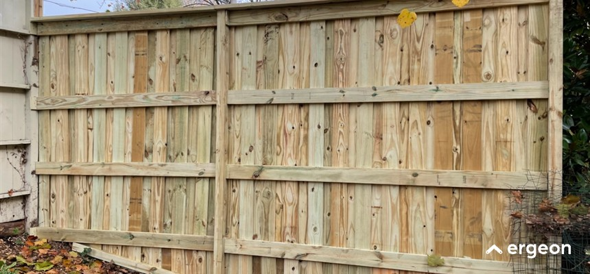Newly Installed Wooden Fence Section in Outdoor Environment This image shows a recently constructed wooden fence section composed of vertical planks supported by horizontal wooden rails. The fence appears to be installed outdoors, likely in a backyard or garden area, with fallen autumn leaves on the ground nearby. The wood used is untreated or lightly treated, showing natural color variations and knots. The fence serves as a privacy barrier and boundary marker, illustrating typical residential fencing construction.