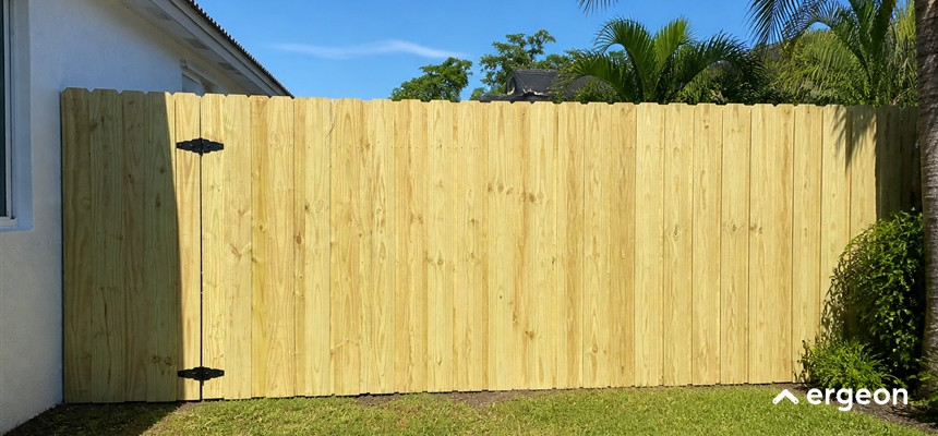 Wooden Privacy Fence with Hinged Gate Installed in Residential Yard This image shows a newly installed wooden privacy fence made of vertical wooden planks with a natural light wood finish. The fence includes a hinged gate on the left side, visible by the black metal hinges fixed to the wood. The fence stands alongside a white residential building wall and encloses a green lawn area. Palm trees and other greenery can be seen in the background under a clear blue sky. The fence is designed to provide privacy and security to a residential property while maintaining an aesthetic appeal.