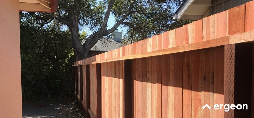 Newly Installed Wooden Fence in Backyard The image shows a recently installed wooden fence made of vertical wooden planks and horizontal support beams. The fence is set alongside a house with a peach-colored exterior wall on the left side and extends into an outdoor backyard area with trees and shrubs visible in the background. The wood appears fresh and untreated, indicating recent construction. The fence provides privacy and security for the property.