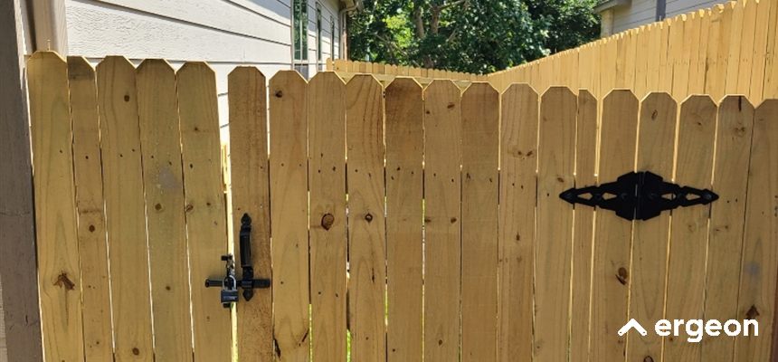 Wooden Privacy Fence Gate with Black Metal Hardware This image depicts a wooden privacy fence gate constructed from vertical wooden planks with a natural finish. The fence panels have a pointed top design and are secured with black metal hardware, including a hinge and a latch system for locking. The fence is positioned between two structures, providing a secure and aesthetically pleasing boundary ideal for residential properties, suitable for outdoor weather conditions such as those in Phoenix.