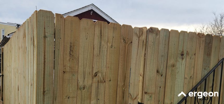 Wooden Privacy Fence Installation in a Pittsburgh Residential Area The image shows a newly installed wooden privacy fence in a residential neighborhood in Pittsburgh. The fence is made of vertically aligned wooden planks with a scalloped top design, providing a secure and private boundary for the property. The background includes parts of neighboring houses, and the fence appears to be freshly constructed, exhibiting clean and uniform wood panels. This photo serves as an example of typical residential fence installation for property demarcation and privacy enhancement.