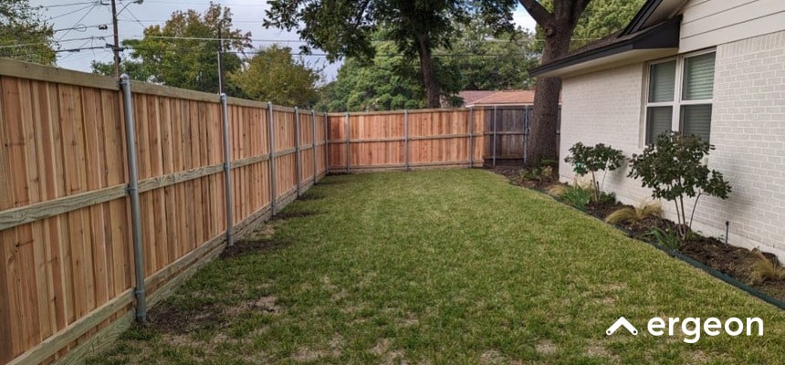 Planned Residential Backyard with Wooden Fence in Plano The image depicts a well-maintained residential backyard enclosed by a newly installed wooden privacy fence. The fence consists of vertical wooden panels reinforced with horizontal support beams and metal posts, providing sturdy boundary delineation. The yard features a green grassy lawn, bordered by a white brick house on the right side and some small garden plants near its foundation. Mature trees and overhead utility lines are visible beyond the backyard fence. This image can be used to demonstrate common homeowner association (HOA) fence styles and regulations in Plano, showcasing acceptable materials and installation standards in residential neighborhoods.