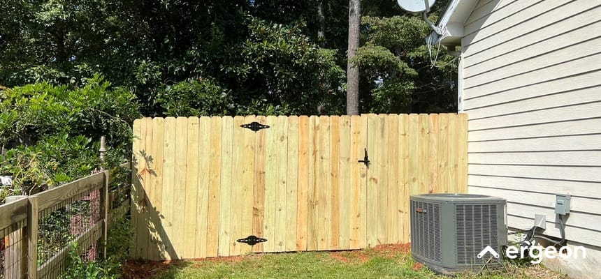 Wooden Fence Gate Installed in Residential Yard in Raleigh This image shows a newly installed wooden fence gate in the backyard of a residential property in Raleigh. The gate is constructed from vertical wooden planks supported by black metal hinges and a latch. It is positioned adjacent to a house with beige siding and near an outdoor air conditioning unit. To the left of the gate, there is an older wire and wooden fence enclosing the yard. The fence gate provides privacy and security while contributing to the outdoor aesthetic.