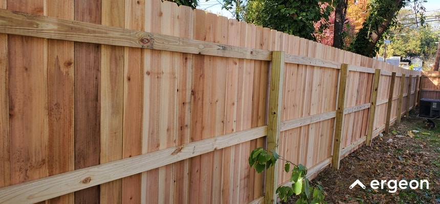 This image depicts a newly installed wooden privacy fence in an outdoor yard setting, likely in New Brunswick. The fence consists of vertical wooden planks secured by horizontal support rails and posts, showcasing a clean and sturdy construction. The background includes trees and foliage, indicating a residential environment. The image serves as a visual example for fencing installation timelines, quality, and materials used in wooden fences.