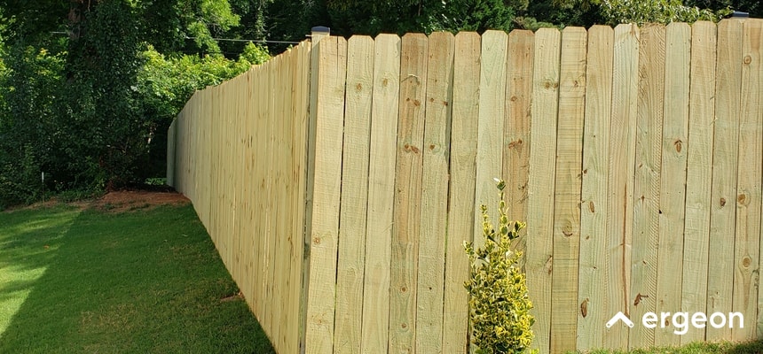 This image features a newly installed wooden privacy fence constructed from closely placed timber planks. The fence encloses a grassy backyard area adjacent to a dense green tree line, providing privacy and boundary definition for the residential property. The fence is tall and solid, designed to obstruct visibility and enhance security. This type of fencing is commonly used in suburban homes for aesthetic appeal and functional privacy.