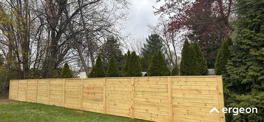 This image shows a long, tall wooden privacy fence made of horizontal planks installed in a suburban yard. The fence is constructed in sections supported by vertical posts, providing a solid barrier for privacy. Behind the fence are tall evergreen trees and leafless deciduous trees, indicating a residential outdoor environment during early spring or late autumn. The cloudy sky suggests overcast weather conditions. The fence serves as a boundary and privacy solution for residential properties.