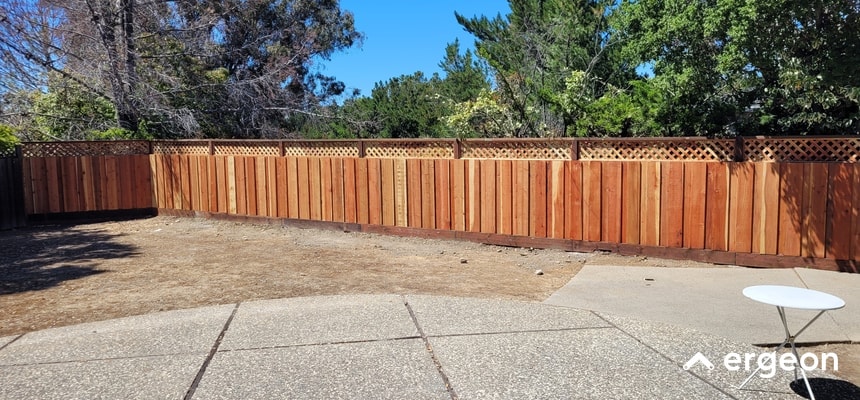 This image shows a newly installed wooden fence in a backyard setting, featuring vertical wooden planks with a lattice design on the upper section. The fence runs along the perimeter of the yard, backed by various green trees and bushes, under a clear blue sky. The foreground includes a concrete patio area with a circular white metal table. The overall setting is indicative of a residential area in Sacramento, and the fence design provides both privacy and an aesthetic appeal suitable for the local weather conditions.
