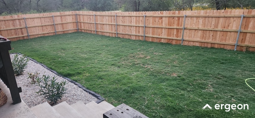 This image shows a freshly installed wooden privacy fence enclosing a rectangular backyard area with well-maintained green grass. The fence is constructed from vertical wooden planks supported by metal posts at regular intervals. In the foreground, there are stone steps leading down to the grassy yard, bordered by a small decorative gravel area with planted shrubs. The scene is set in a suburban environment with trees visible beyond the fence, providing a private and natural outdoor space. The image can be used for illustrating fencing projects, landscaping ideas, or property boundary installations.