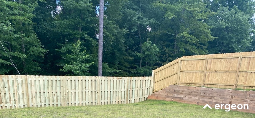 This image shows a recently installed wooden privacy fence surrounding a backyard that borders a wooded area. The fence is constructed from vertical wooden planks with horizontal support beams. The left side features a straight section of fence, while the right side shows a gated corner with steps leading up a slight slope, secured by the fence. The surrounding area includes green grass and dense trees, highlighting the fence's role in providing privacy and security while blending with the natural environment.