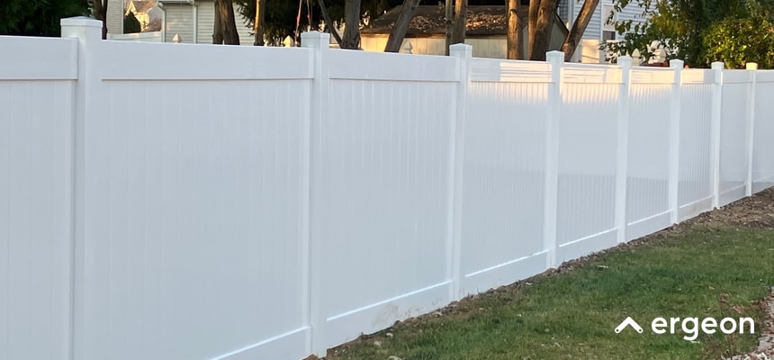 This image depicts a long stretch of white vinyl privacy fencing installed along the edge of a grassy yard in a suburban neighborhood. The fence panels are solid, with vertical slats and reinforcing horizontal rails, designed for privacy and security. Trees and other residential houses are visible in the background, indicating a typical suburban setting. The fence is well-maintained, clean, and uniformly installed, making it a suitable example for documentation on residential fencing options, installation standards, and neighborhood property boundary delineation.