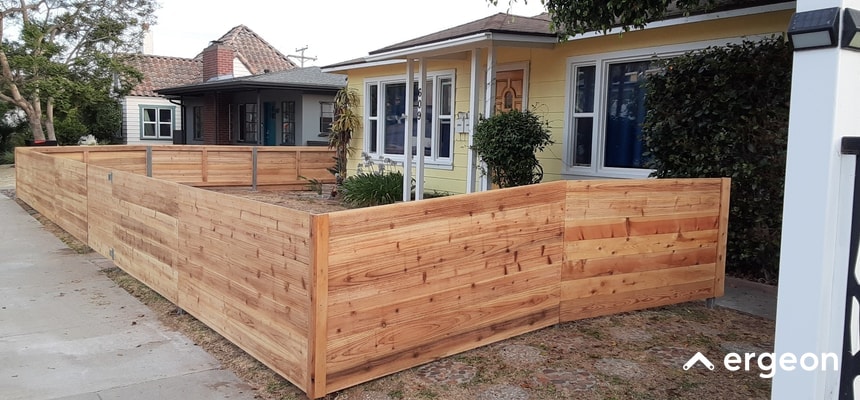 The image shows a modern, well-constructed cedar wood fence enclosing the perimeter of a residential front yard. The fence is made from horizontal wooden planks with a natural finish, providing a clean and contemporary aesthetic. It is of medium height, offering privacy while still maintaining an open and inviting appearance. The fence is complemented by the surrounding homes and landscaping, demonstrating an effective use of wooden fencing for residential boundary demarcation and aesthetic enhancement.