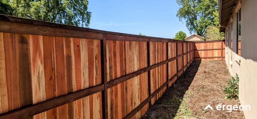 The image shows a newly installed cedar wooden fence along the sideyard of a residential property. The fence consists of vertically aligned cedar planks secured between horizontal support beams, providing privacy and security. The fence runs parallel to the exterior wall of the house, enclosing the side yard area, with a clear blue sky and some greenery visible in the background. This type of fence is typically used for enhancing property boundaries and outdoor aesthetics.