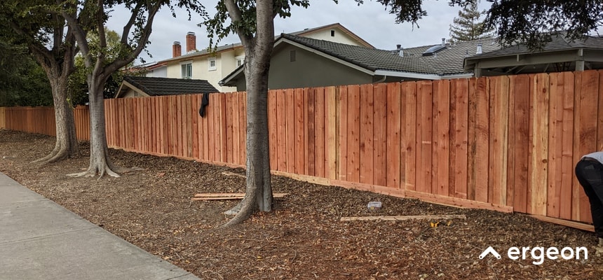 The image depicts a newly constructed wooden privacy fence running along a residential property boundary in a suburban neighborhood. The fence is made of vertical wooden panels and extends along the edge of a landscaped yard with a sidewalk adjacent to it. Mature trees are spaced intermittently along the length of the fence, which provides both privacy and aesthetic appeal to the properties behind it. The photo highlights the fence's height, uniform construction, and natural wood finish, illustrating an effective and durable fencing solution for residential outdoor spaces.