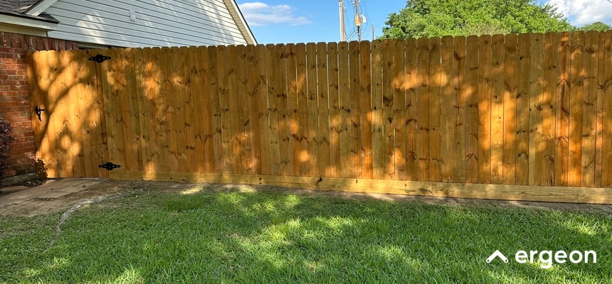 This image shows a newly installed wooden privacy fence made of vertical wooden planks with a natural finish. The fence is positioned beside a house and includes a gate with black metal hinges and latch hardware. The fence is designed to provide privacy and security for the residential property. The surrounding area features a well-maintained grassy lawn and clear blue sky, indicating an outdoor setting suitable for protecting yard boundaries from external view and enhancing home security.